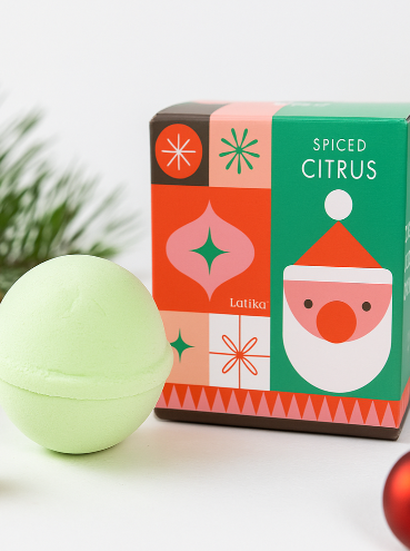 Green bath bomb next to a colorful box labeled 'Spiced Citrus' with Christmas decorations in the background.